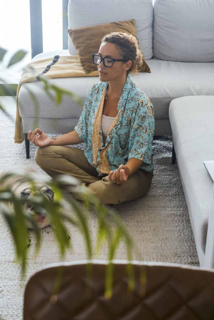 A woman with a moderate complexion and glasses meditating peacefully on the floor beside her couch