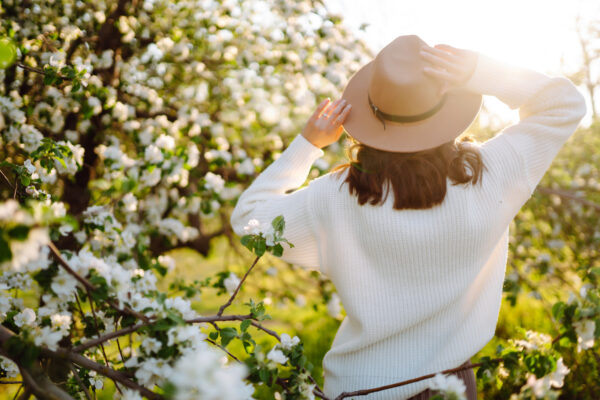 Portrait of Beautiful Woman In hat posing near flowering. Smiling woman enjoying smell of flowers.