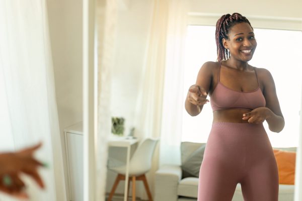 Young fit black woman in sportswear dancing in front of mirror while having domestic fitness training and smiling. Sporty lady working out indoors and having fun